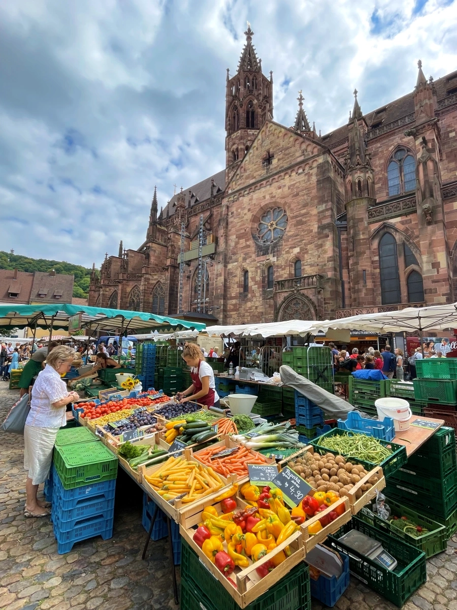 Mujer comprando verduras en puesto del Münstermarkt en la plaza de la catedral de Friburgo, con la torre gótica de 116 metros al fondo