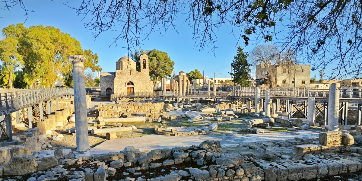 Vista de la iglesia de Agia Kyriaki Chrysopolitissa rodeada de columnas y ruinas arqueológicas en Pafos, Chipre, con pasarelas de madera y árboles al fondo