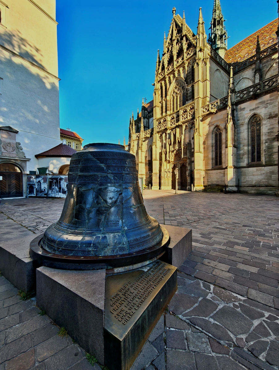 Primer plano de la campana original de San Urbano, fundida en 1557, situada en la plaza frente a la Catedral de Santa Isabel en Košice, Eslovaquia, con detalles góticos de la catedral al fondo y placa conmemorativa en la base.