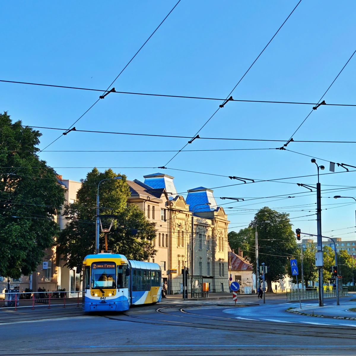Tranvía azul y amarillo circulando por una avenida de Košice, Eslovaquia, con cables eléctricos visibles sobre la calle y edificios históricos al fondo, en una mañana despejada. Transporte público sostenible en Košice.