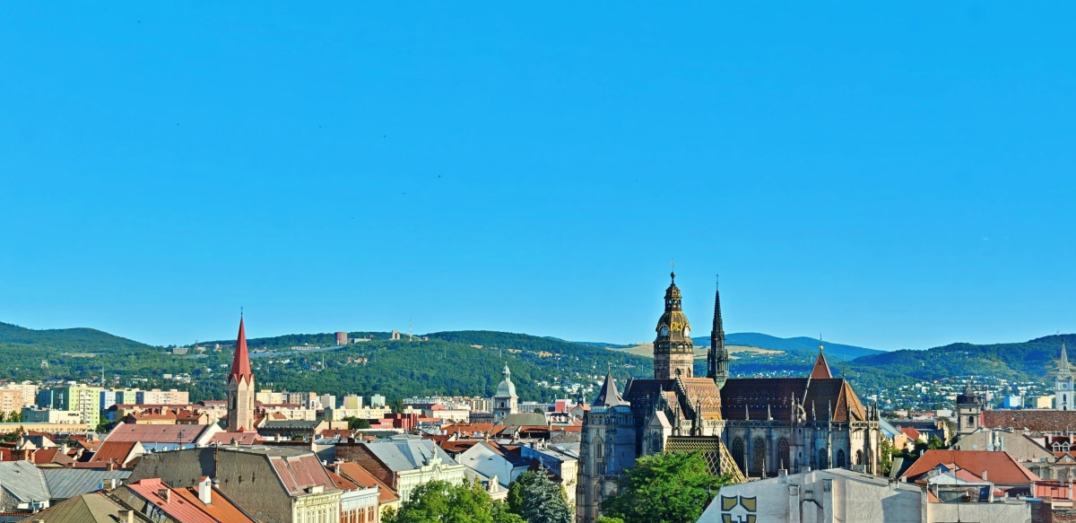 Vista panorámica del centro histórico de Košice, Eslovaquia, destacando la Catedral de Santa Isabel con su tejado colorido y torres góticas, rodeada de tejados rojizos y colinas verdes bajo un cielo azul claro.