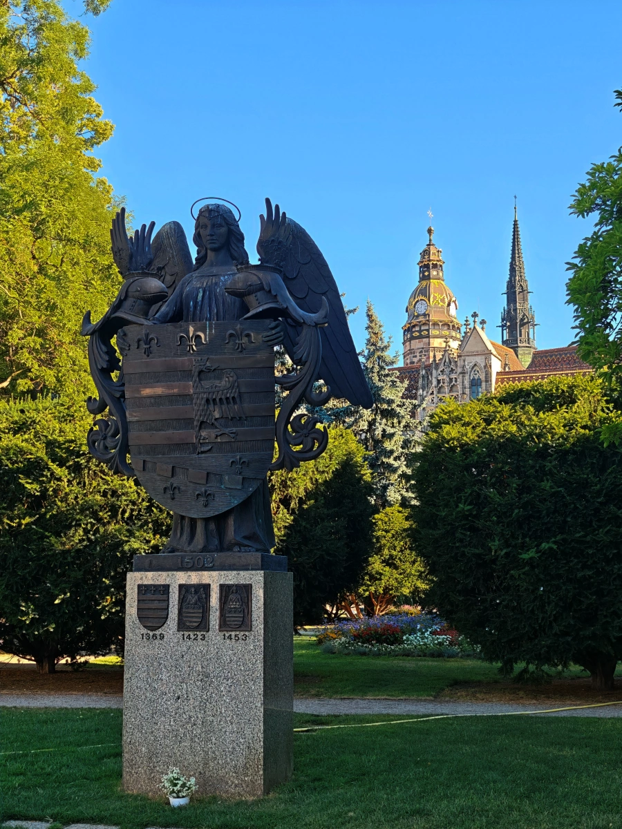 Monumento del ángel alado sosteniendo el escudo de armas de Košice, con la Catedral de Santa Isabel al fondo y jardines verdes, símbolo histórico de la ciudad eslovaca.