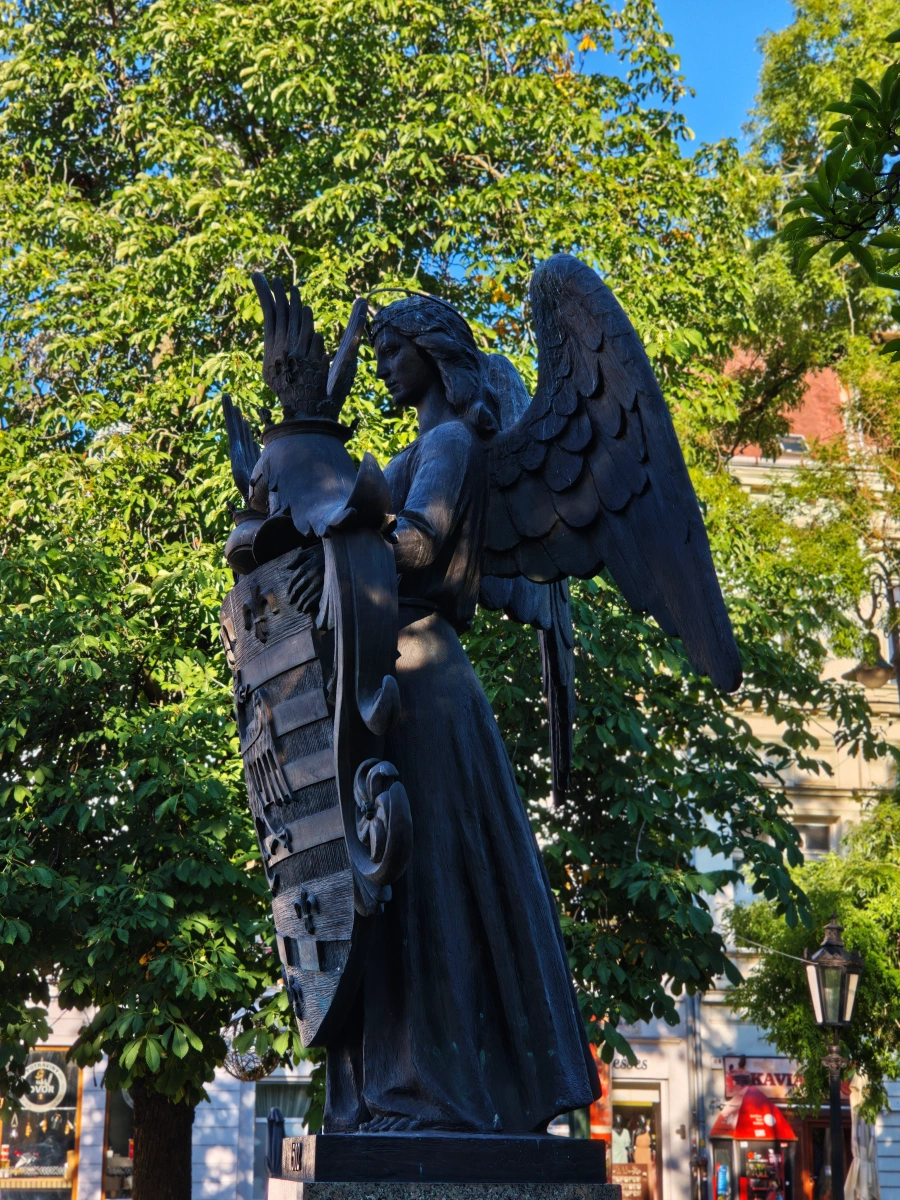 Vista lateral de la escultura de un ángel alado sosteniendo el escudo de armas de Košice, rodeada de árboles y vegetación en el centro histórico.