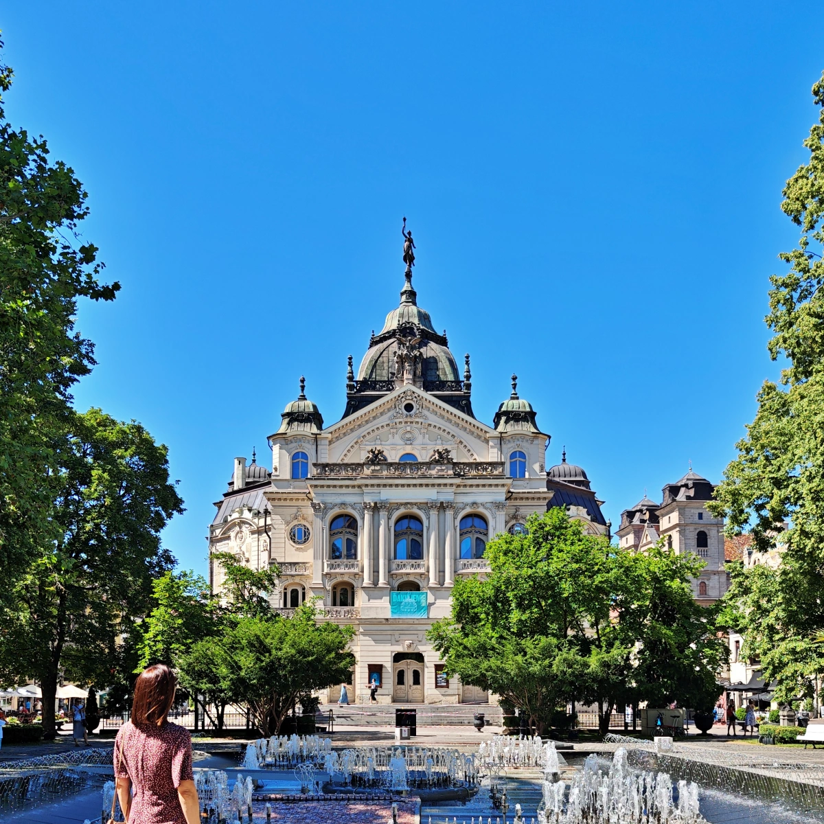 ista frontal del Teatro Estatal de Košice, Eslovaquia, con su fachada neobarroca, cúpula central, esculturas ornamentales y una fuente rodeada de árboles bajo un cielo azul.