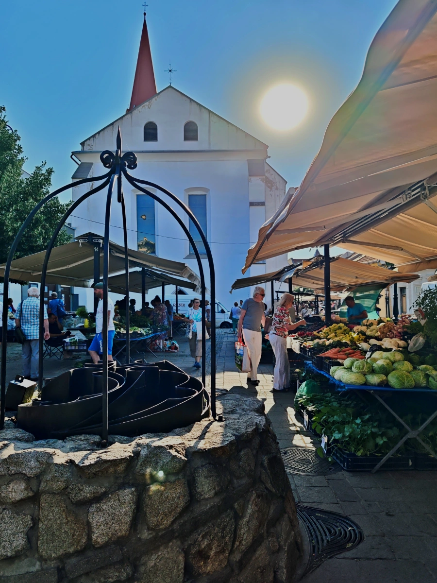 Pozo de piedra en primer plano y puestos de verduras frescas en el mercado de la Plaza Dominicana de Košice, con la iglesia al fondo y el sol brillante.