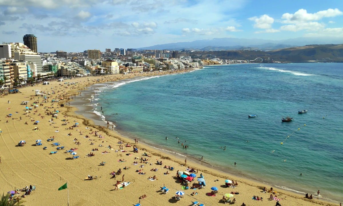 Imagen panorámica de la Playa de Las Canteras tomada desde La Puntilla, con arena dorada, bañistas y la ciudad al fondo bajo cielo azul.