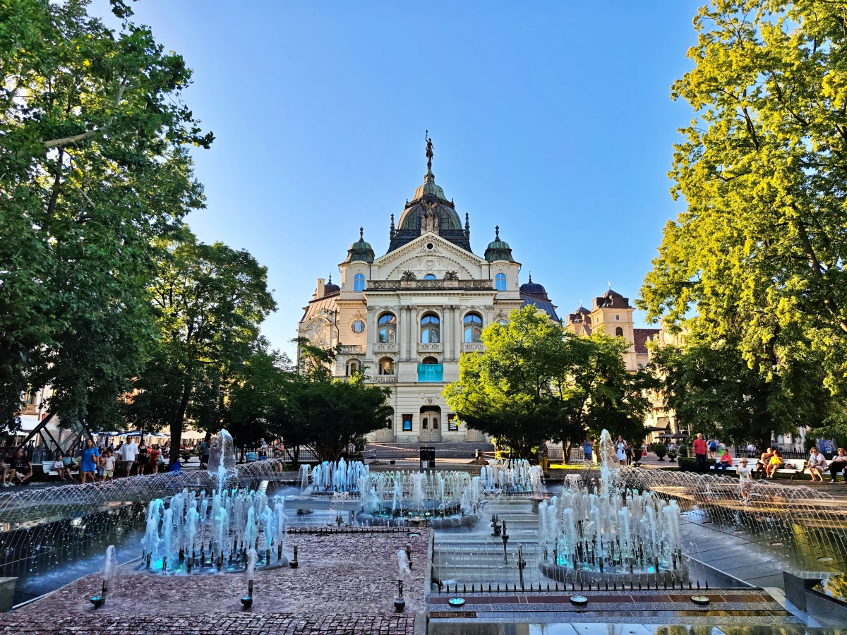 Fuente Musical de Košice iluminada al atardecer, con chorros de agua sincronizados frente al Teatro Estatal, rodeada de árboles y visitantes en la calle Hlavná, centro histórico de Košice, Eslovaquia.