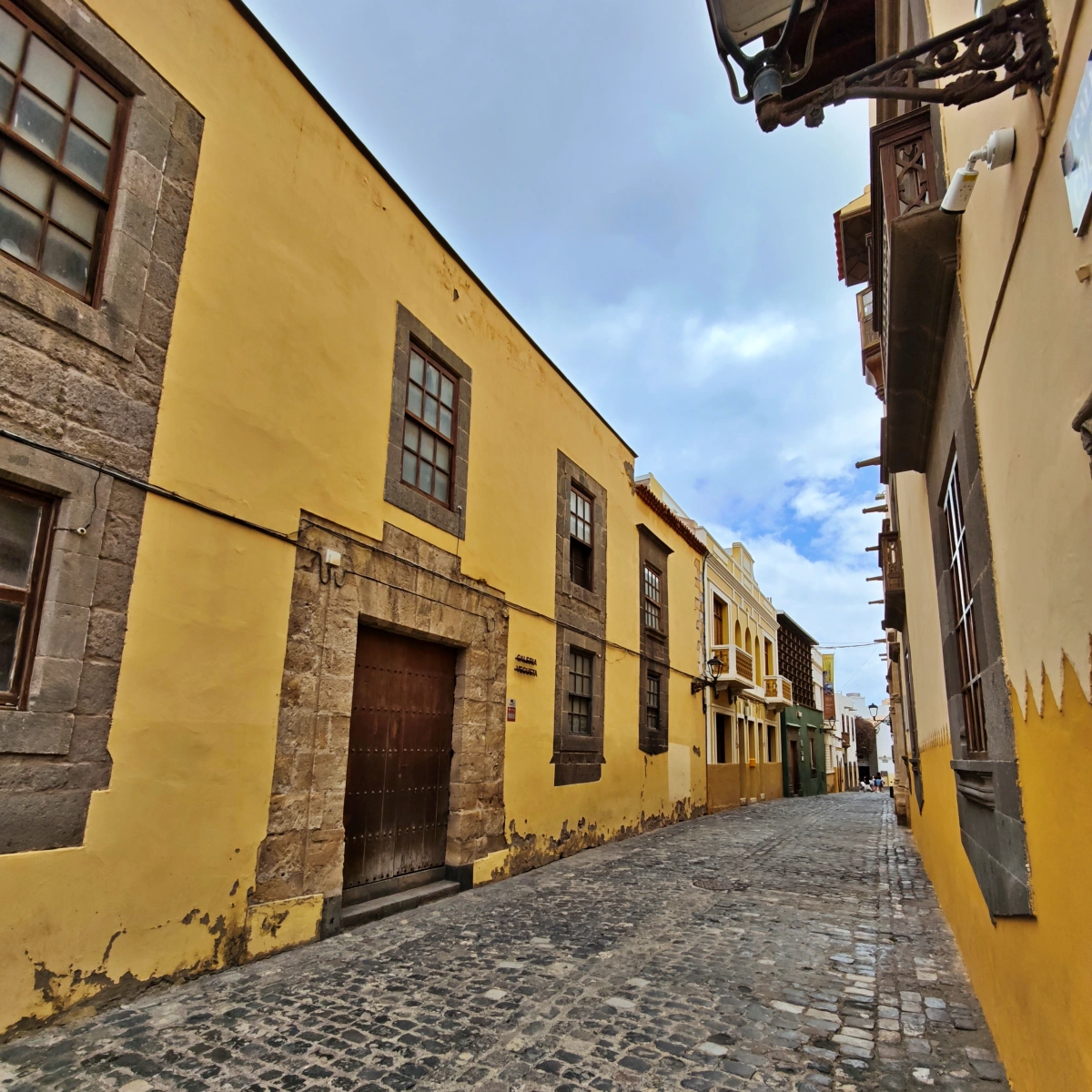 Calle estrecha y empedrada del barrio de Vegueta con casas históricas de fachadas amarillas y ventanas de madera bajo cielo parcialmente nublado.