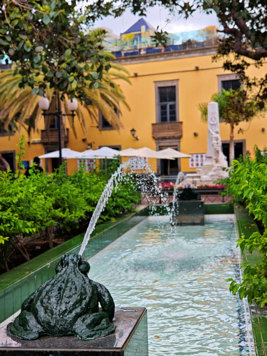 Detalle artístico de la fuente con esculturas de ranas en la Plaza de las Ranas, rodeada de vegetación y arquitectura histórica en Las Palmas.