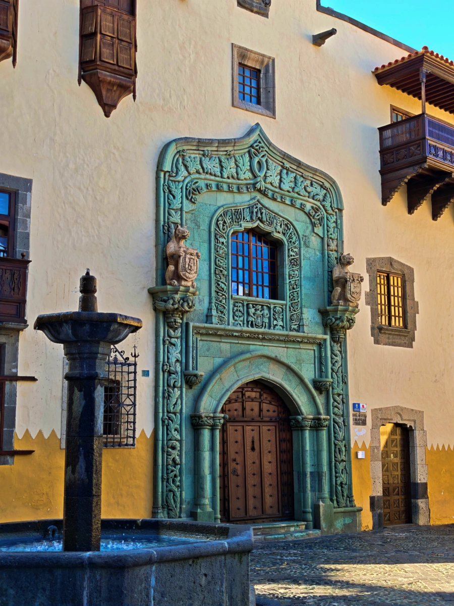 Fachada principal de la Casa de Colón en Las Palmas de Gran Canaria con portada de piedra verde, balcones de madera y fuente al frente.