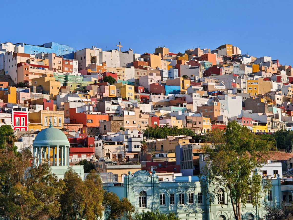 Fotografía panorámica del Paraninfo de la Universidad de Las Palmas de Gran Canaria, con su emblemática cúpula verde en primer plano y, al fondo, el Barrio de San Juan con sus casas multicolores en la colina bajo el cielo azul.