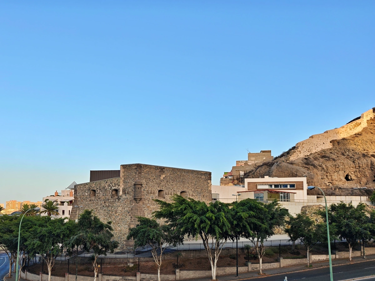 Vista del Castillo de Mata en Las Palmas de Gran Canaria, una fortificación histórica de piedra rodeada de árboles y colina al fondo bajo un cielo despejado.