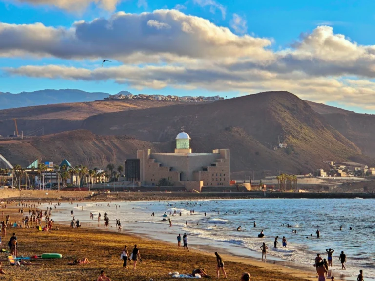 Vista amplia de la Playa de Las Canteras en Las Palmas de Gran Canaria con personas disfrutando del mar, el Auditorio Alfredo Kraus en el fondo y el Barrio de Los Giles en la ladera.