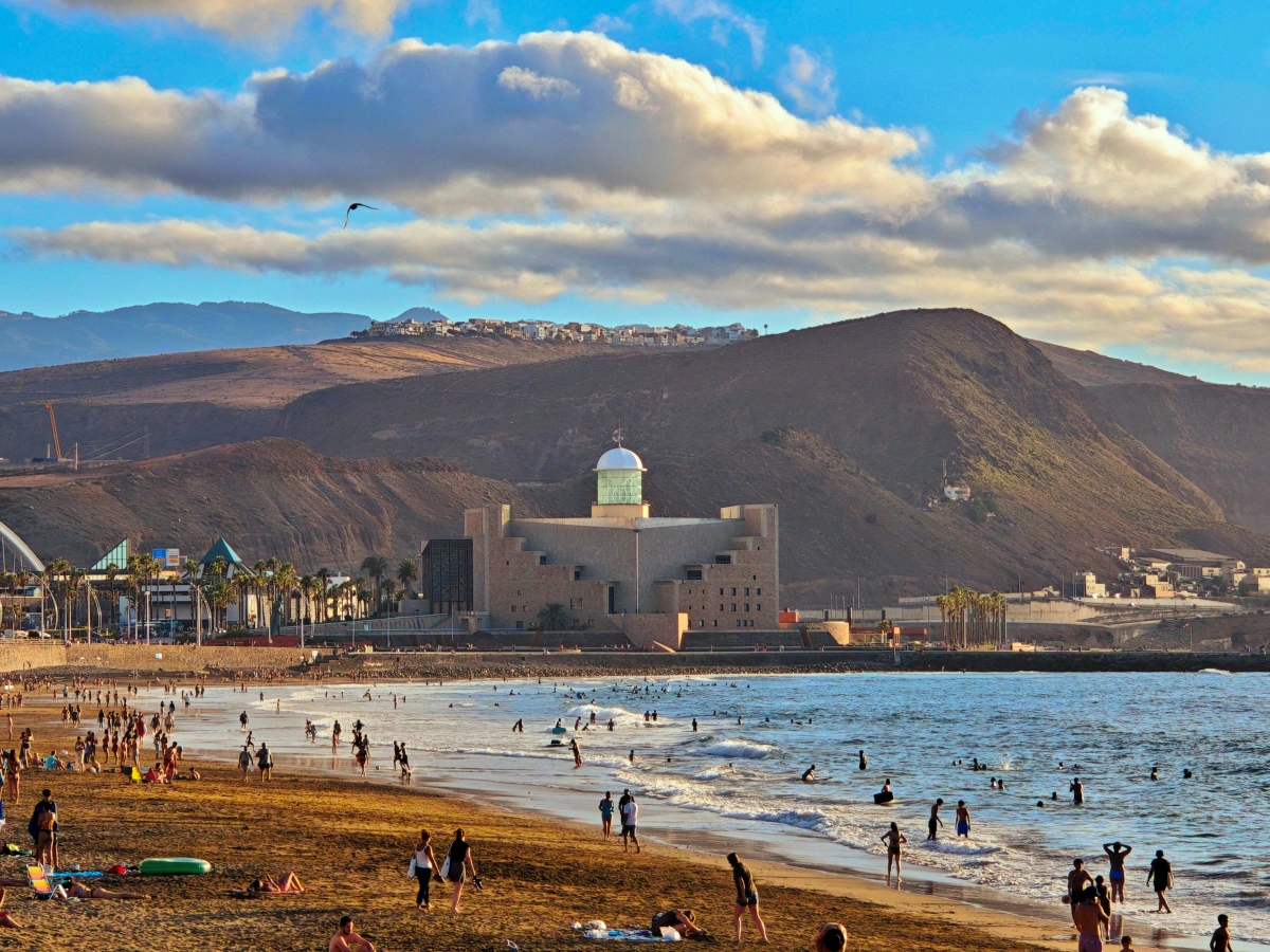 Vista amplia de la Playa de Las Canteras en Las Palmas de Gran Canaria con personas disfrutando del mar, el Auditorio Alfredo Kraus en el fondo y el Barrio de Los Giles en la ladera.