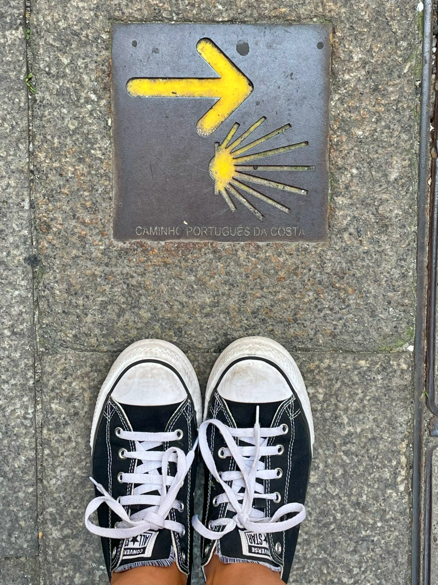 Mosaico en el suelo con flecha amarilla y concha del Camino Portugués de la Costa junto a zapatillas