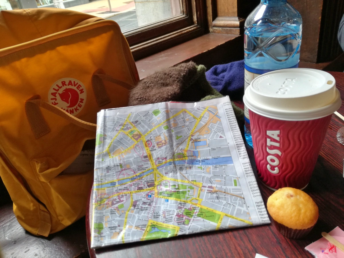 Mochila amarilla Fjällräven, mapa turístico, café Costa y muffin sobre mesa de cafetería junto a ventana