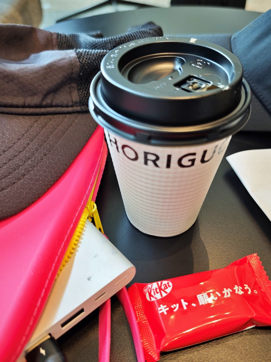 Gorra negra, estuche rosa, batería portátil, café para llevar y KitKat japonés sobre mesa negra