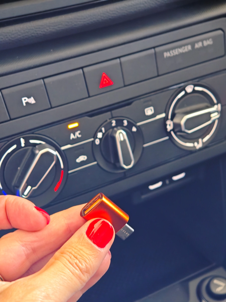 Mano con uñas rojas sosteniendo un adaptador USB-C frente a la consola de climatización de un coche