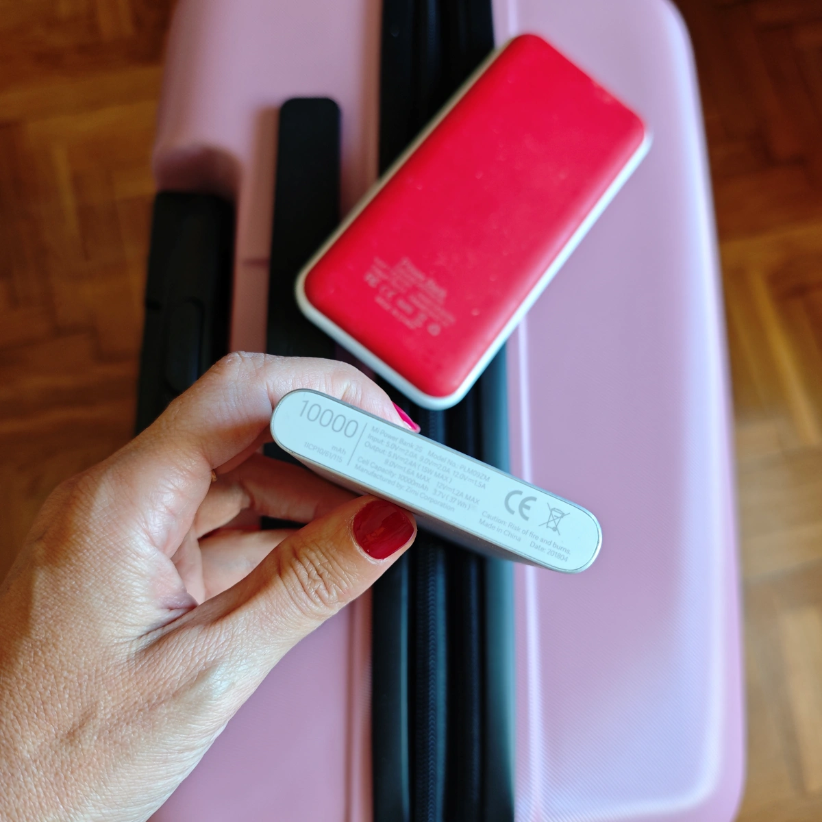 Mano con uñas rojas sujetando batería externa de 10.000 mAh sobre una maleta rosa