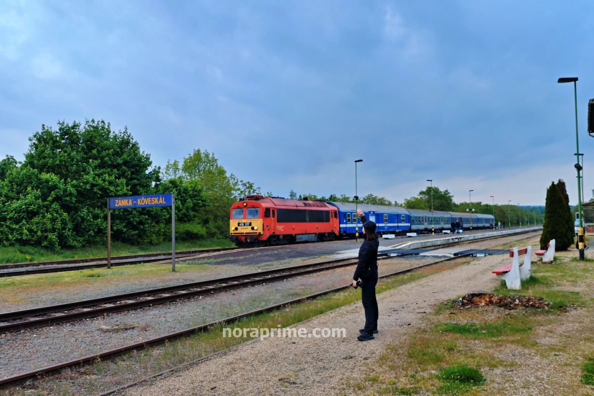Locomotora naranja y vagones azules de MÁV en la estación Zánka-Köveskál junto a señales y bancos al aire libre, Hungría