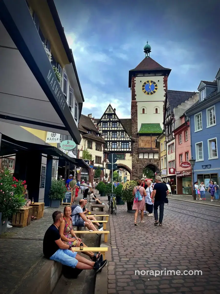 Gente disfrutando de bebidas en terraza junto al Bächle cerca del Schwabentor en Friburgo, escena típica de la vida local