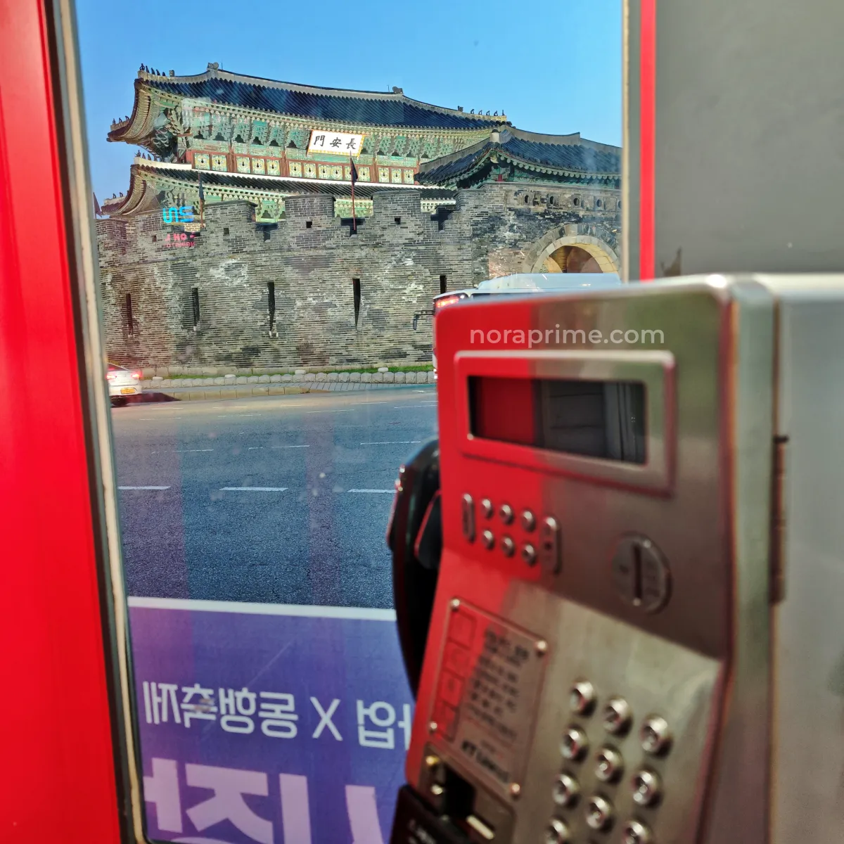 Teléfono público dentro de una cabina roja en Suwon, Corea del Sur, con la puerta Janganmun (장안문) de la fortaleza Hwaseong al fondo, muralla histórica iluminada al atardecer.
