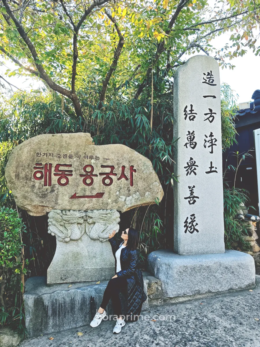 Señal de piedra en la entrada de Haedong Yonggungsa (해동용궁사) en Busan, Corea del Sur, con inscripción en coreano y un monolito con caligrafía, rodeada de bambú y árboles, junto al acceso al templo costero.