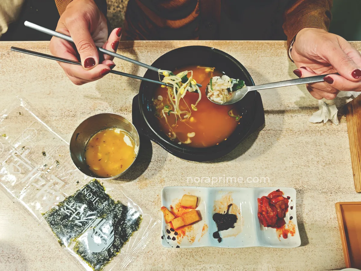 Persona comiendo kongnamul gukbap, sopa coreana con brotes de soja y arroz en cuenco negro, acompañada de banchan como kimchi y kkakdugi, y paquete de alga gim sobre la mesa.