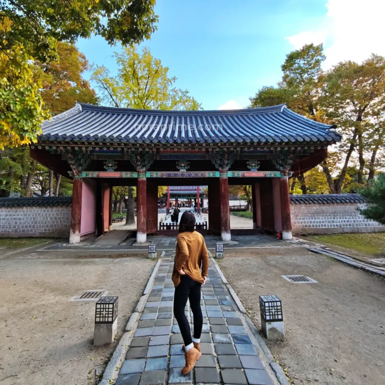 Viajera caminando hacia una puerta tradicional coreana en Jeonju Hanok Village rodeada de árboles en otoño