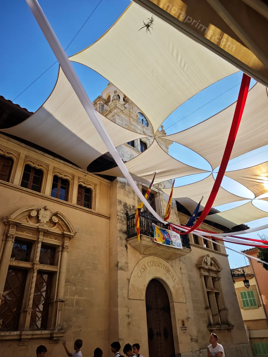 Casa Consistorial de Alcúdia en el casco histórico, con torre del reloj, balcones con banderas y toldos blancos y rojos sobre la plaza llena de gente.