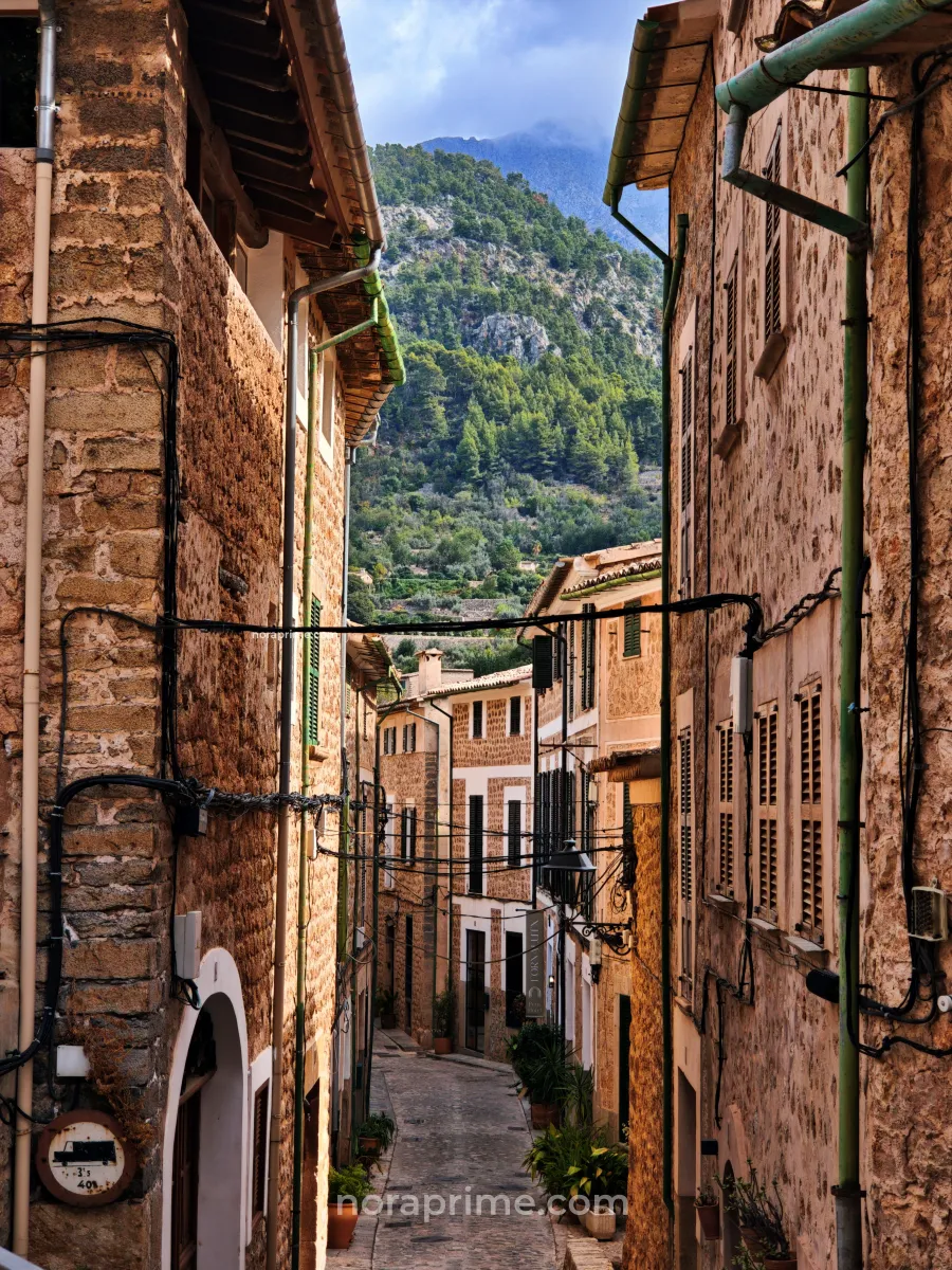 Calle empedrada de Fornalutx entre casas de piedra y persianas mallorquinas, con montañas de la Serra de Tramuntana al fondo en Mallorca.