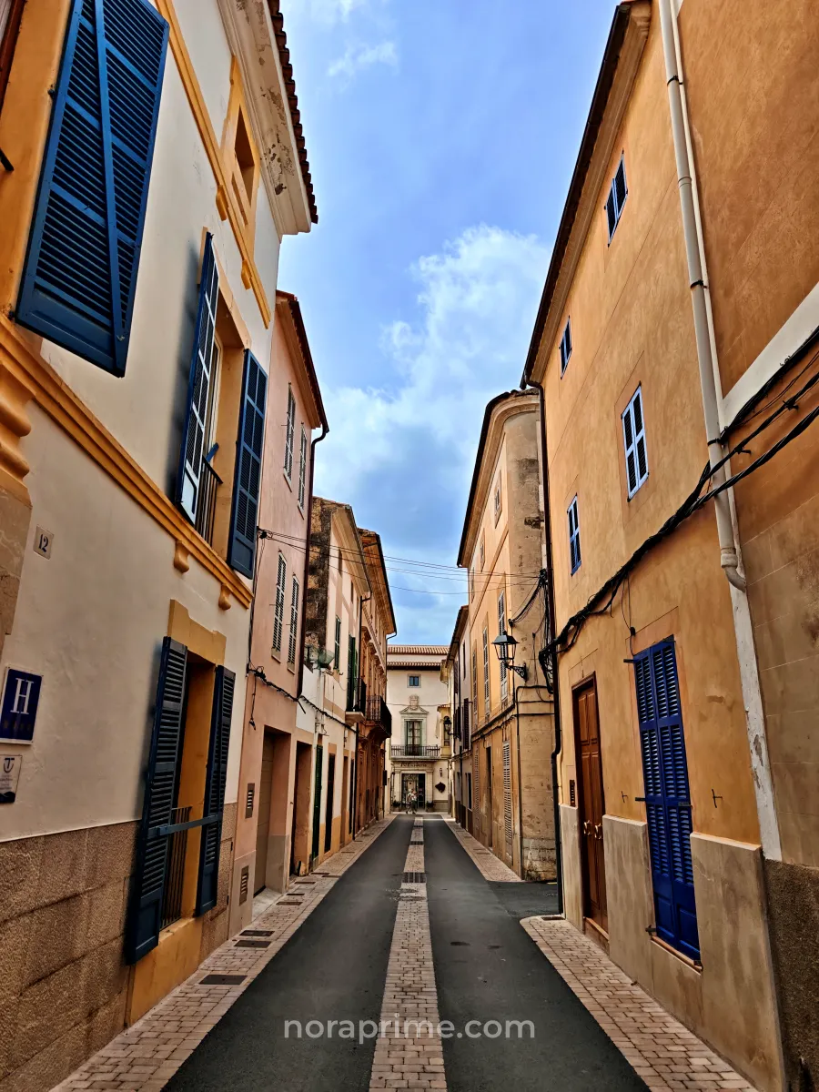 Calle estrecha y empedrada en el centro histórico de Artà, Mallorca, con fachadas ocre y contraventanas azules típicas de los pueblos bonitos del Mediterráneo.