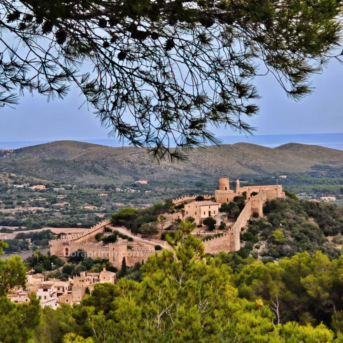 Panorámica del Castillo de Capdepera en Mallorca, recinto amurallado medieval del siglo XIV situado en lo alto de una colina, rodeado de bosques y montañas, con vistas al pueblo de Capdepera y al mar Mediterráneo al fondo.