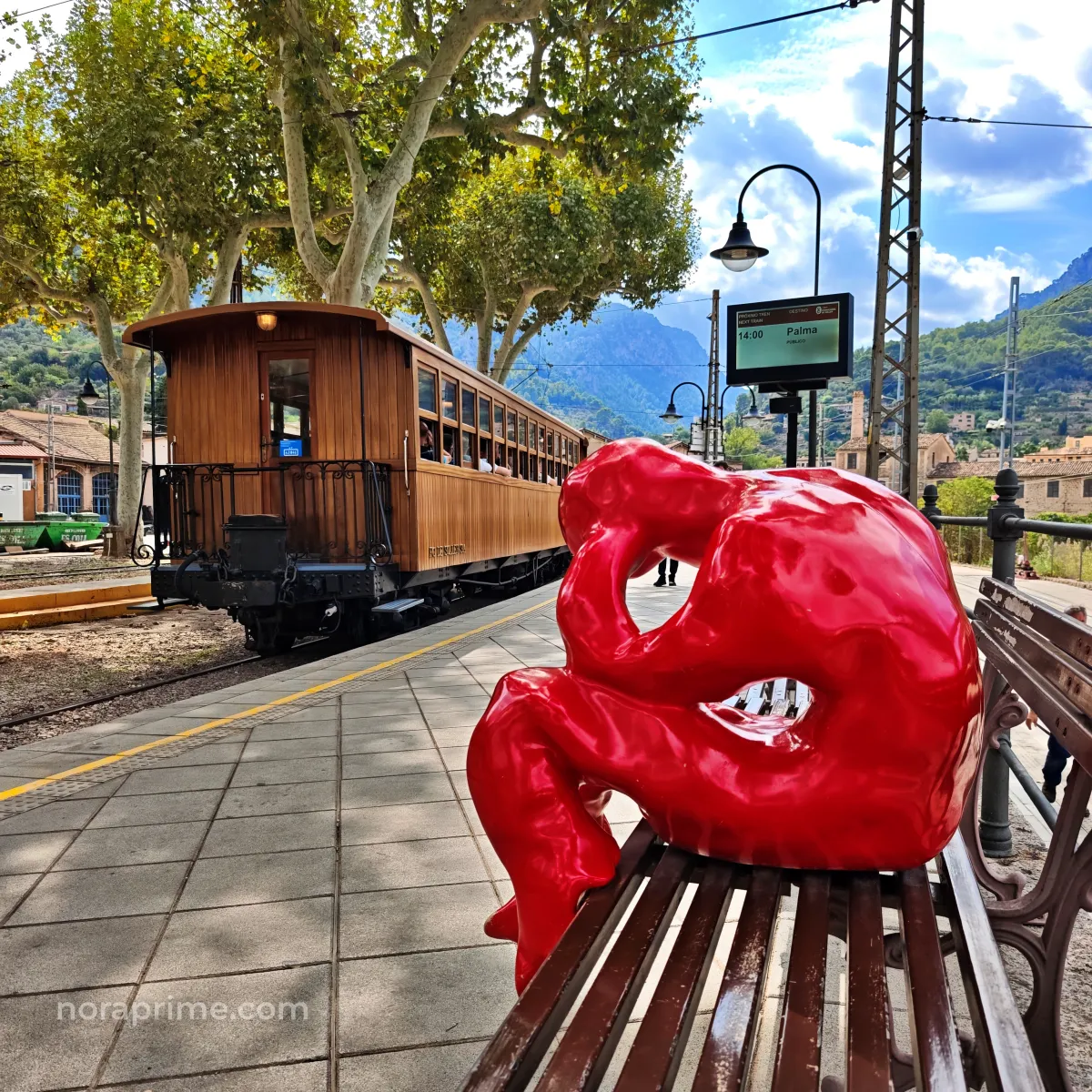 Escultura roja Dreamers en la estación del tren de Sóller, con vagón de madera al fondo y montañas de la Serra de Tramuntana en Mallorca.