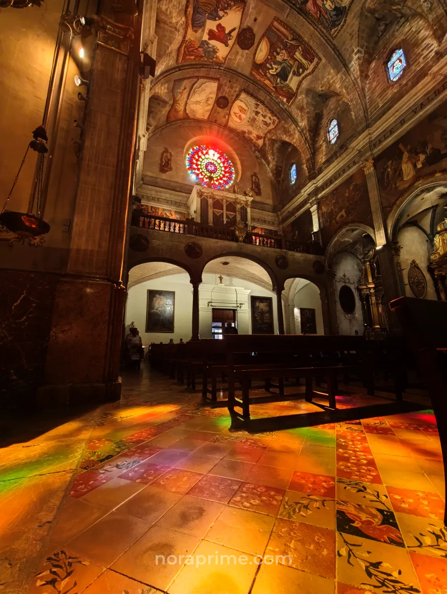 Luz de colores entrando por el rosetón de la iglesia Mare de Déu dels Àngels en Pollença, iluminando el suelo de baldosas y las naves barrocas del templo.