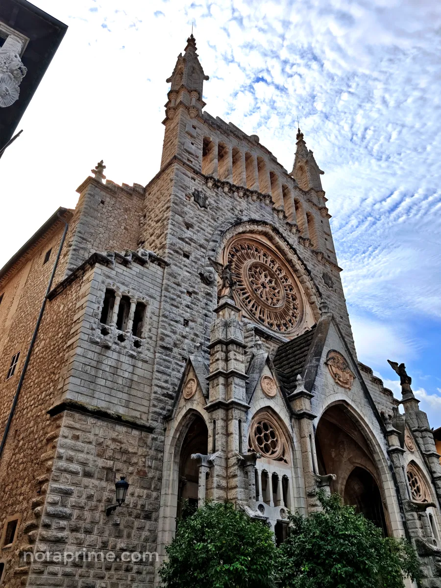 Vista en contrapicado de la fachada modernista de la iglesia de Sant Bartomeu de Sóller, con gran rosetón y torre escalonada, bajo un cielo parcialmente nublado en Mallorca.