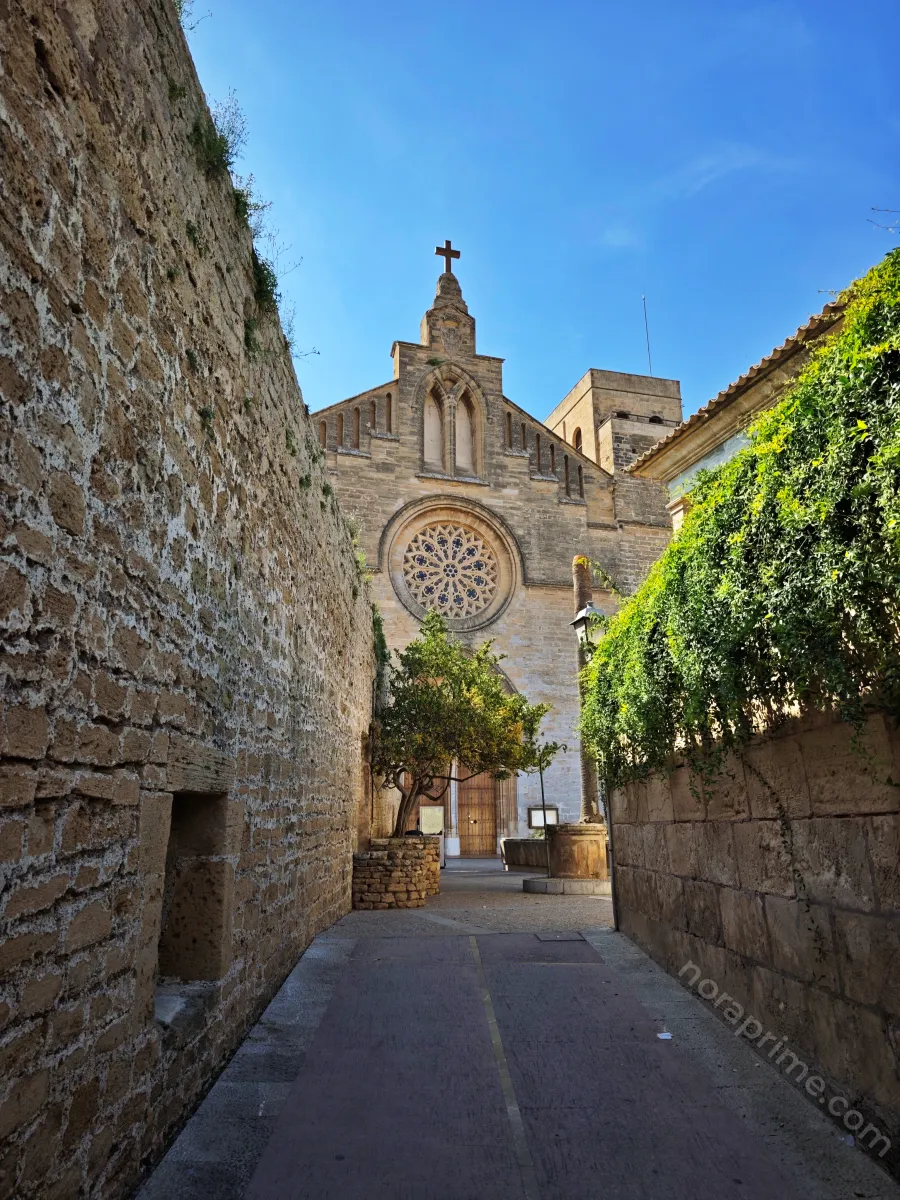 Calle estrecha que conduce a la iglesia de Sant Jaume de Alcúdia, con murallas de piedra a ambos lados y fachada gótica con rosetón y cruz bajo el cielo azul.