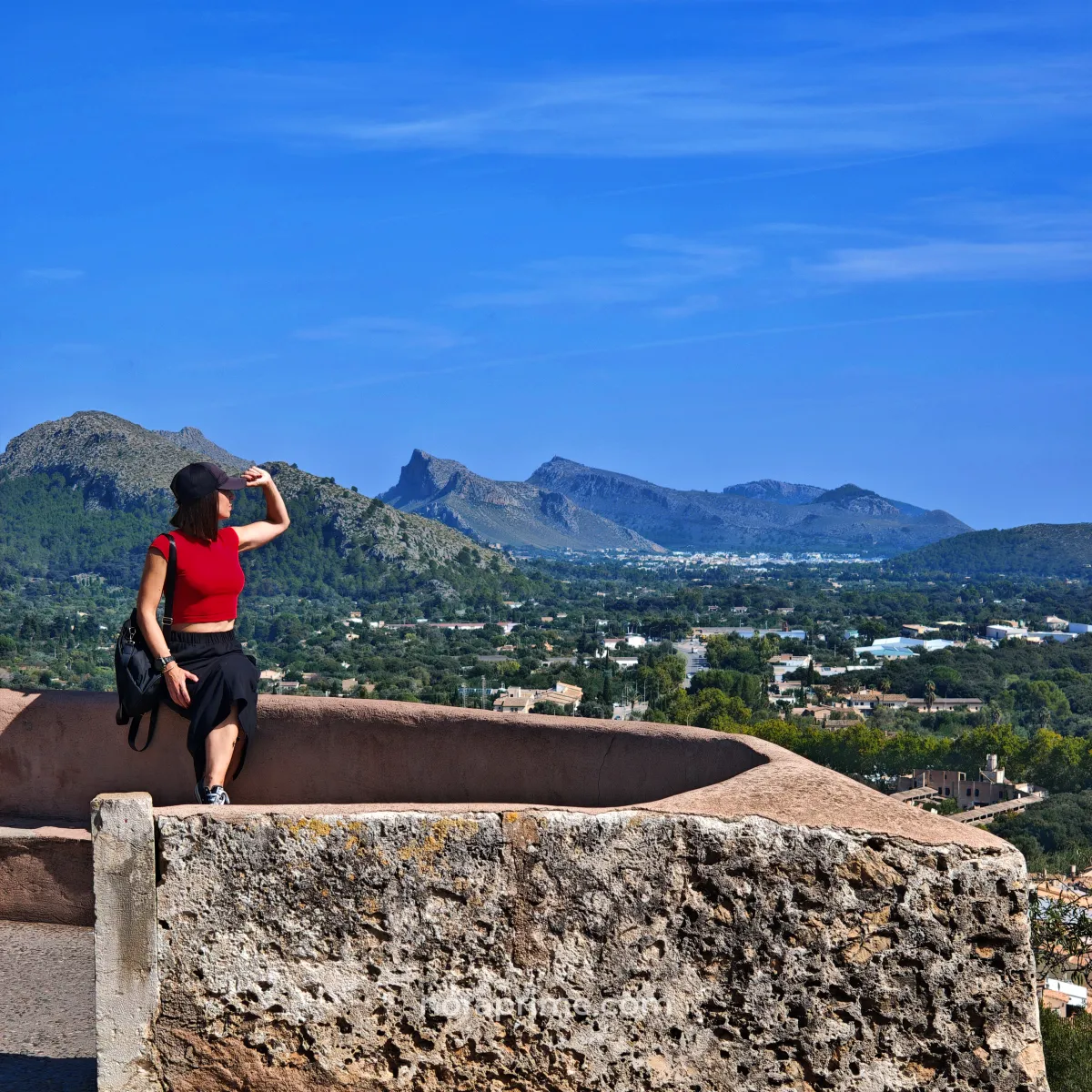 Panorámica desde el mirador del Calvari de Pollença, con una mujer sentada en el muro y las montañas y pueblos del norte de Mallorca al fondo.