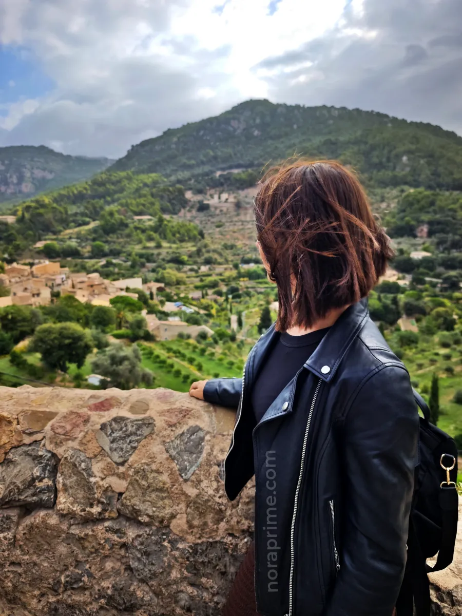 Mujer apoyada en un muro de piedra en un mirador de Valldemossa, con vistas al pueblo, terrazas de cultivo y montañas verdes de la Serra de Tramuntana en Mallorca.