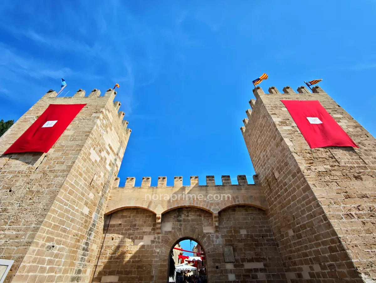 Vista en contrapicado de la Porta de Mallorca en Alcúdia, muralla medieval de piedra con dos torres almenadas y banderas, entrada al casco antiguo de la localidad en el norte de Mallorca.