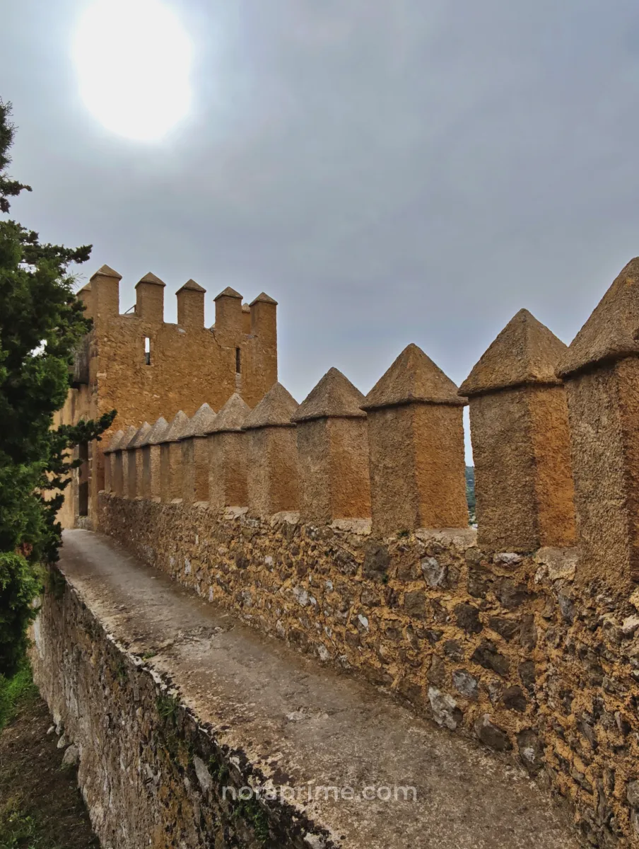 Camino estrecho sobre las murallas de piedra del Santuario de Sant Salvador en Artà, Mallorca, con torre fortificada y vegetación mediterránea alrededor.