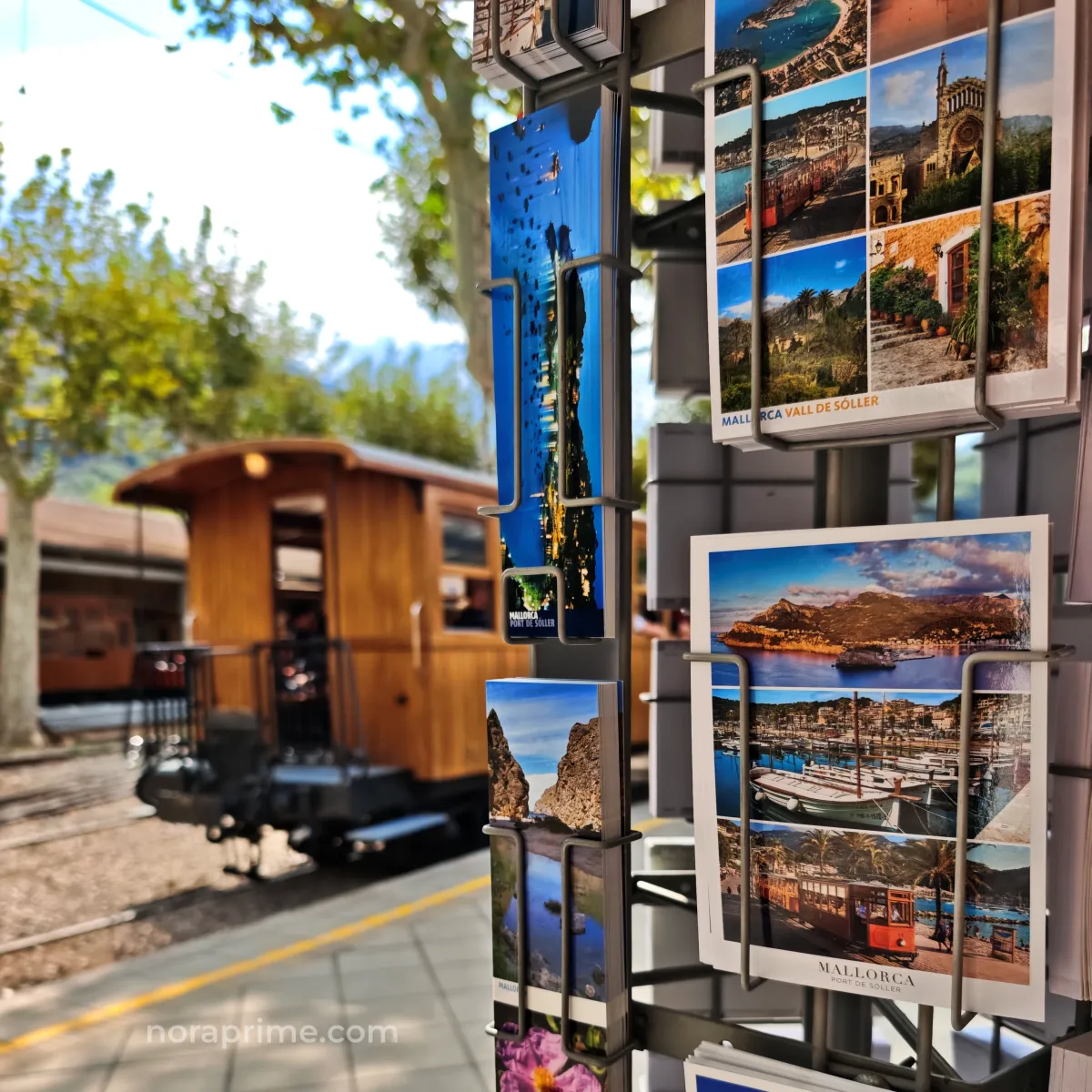 Detalle de postales turísticas de Sóller y Port de Sóller en un soporte metálico, con el tren de madera desenfocado al fondo en la estación de Mallorca.
