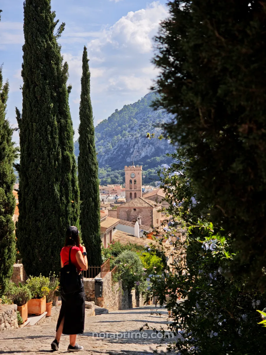 Mujer caminando por la subida al Calvari de Pollença, entre cipreses y casas de piedra, con el campanario del pueblo y la Serra de Tramuntana al fondo en Mallorca.