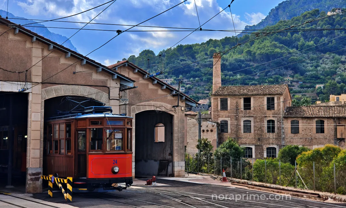 Tranvía de madera de Sóller frente a las cocheras y edificios industriales históricos, rodeado de montañas verdes en el valle de Sóller, Mallorca.